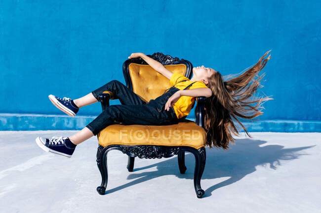 Side view of teen hipster sitting on chair and throwing hair on blue background — Stock Photo