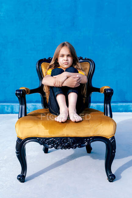 Sad teenager with long hair and in hipster clothes sitting on chair and embracing knees while looking at camera on blue background — Stock Photo