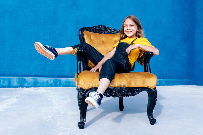 Веселий підліток хіпстер сидить на стільці на синьому фоні, дивлячись далеко — стокове фото