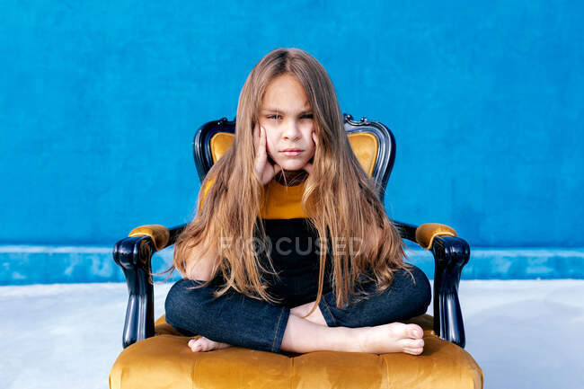Trauriger Teenager mit langen Haaren und in Hipster-Klamotten, der mit gekreuzten Beinen auf einem Stuhl sitzt, während er in die Kamera auf blauem Hintergrund blickt — Stockfoto