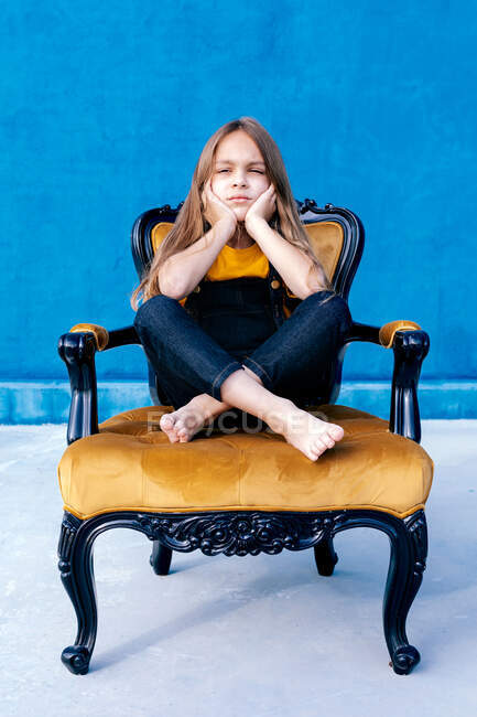Сумний підліток з довгим волоссям і в хіпстерському одязі, сидячи на стільці з перехрещеними ногами, дивлячись на синій фон — стокове фото