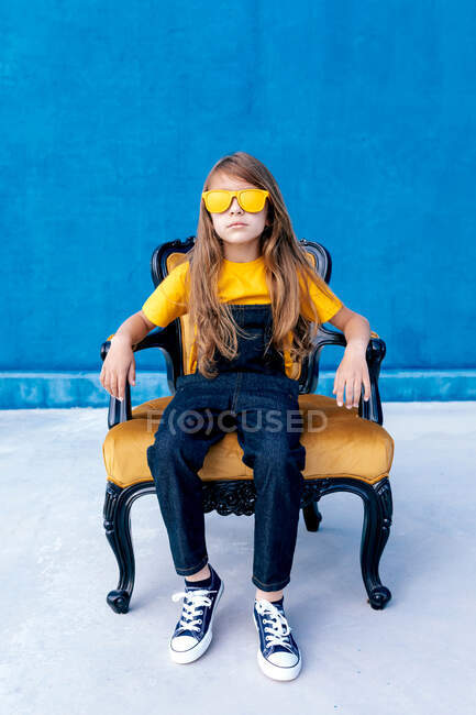 Thoughtful teen hipster sitting on chair with trendy yellow sunglasses on blue background looking at camera — Stock Photo