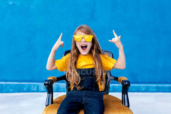 Adolescent expressif aux cheveux longs et aux lunettes de soleil jaunes à la mode montrant un signe rock et criant sur fond bleu — Photo de stock