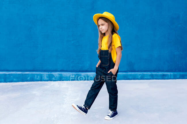 Cool thoughtful teenage hipster in overalls and yellow hat walking with hands in pockets on background of blue wall — Stock Photo