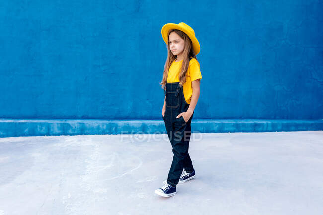 Cool adolescent réfléchi hipster en salopette et chapeau jaune marche avec les mains dans les poches sur fond de mur bleu — Photo de stock
