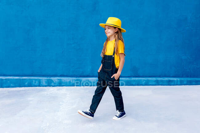 Cool thoughtful teenage hipster in overalls and yellow hat walking with hands in pockets on background of blue wall — Stock Photo