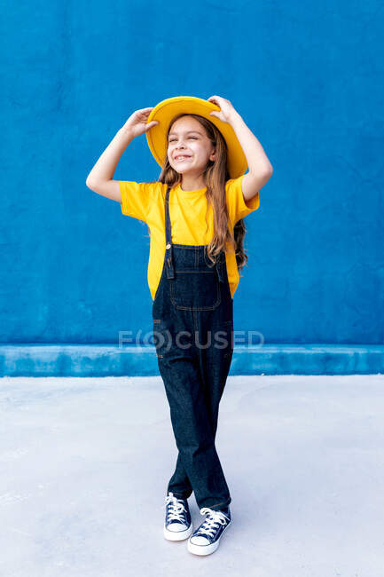 Cool cheerful teenage hipster in overalls and yellow hat standing looking away on background of blue wall — Stock Photo