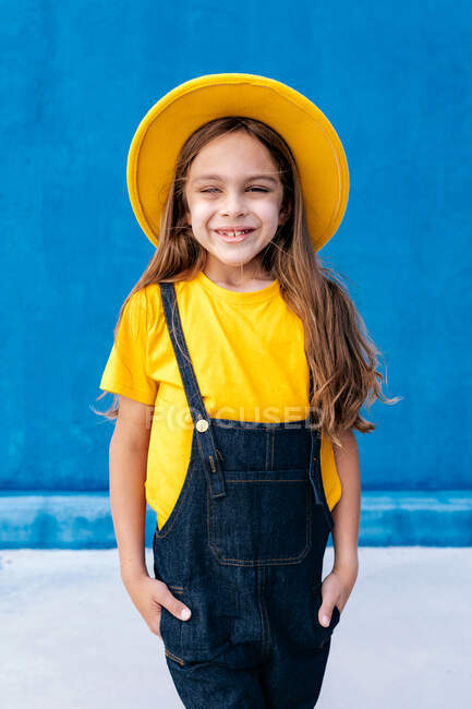 Coole, fröhliche Teenager-Hipster in Overalls und gelbem Hut, die Hände in Taschen auf blauem Wandhintergrund und in die Kamera blickend — Stockfoto