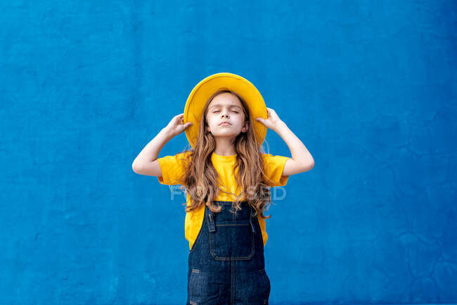 Cool hipster adolescente reflexivo en overol y sombrero amarillo de pie con los ojos cerrados en el fondo de la pared azul - foto de stock