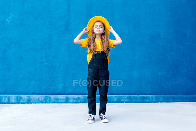 Cool thoughtful teenage hipster in overalls and yellow hat standing with eyes closed on background of blue wall — Stock Photo