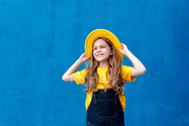 Cool adolescent gai hipster en salopette et chapeau jaune debout regardant loin sur fond de mur bleu — Photo de stock