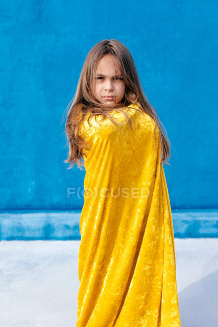Serious teenager with long hair wrapped in yellow cloak standing on blue background and looking at camera — Stock Photo