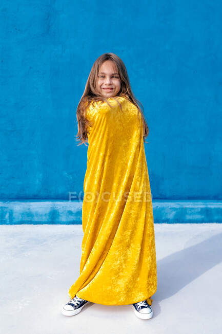 Adolescente feliz com cabelos longos envolto em capa amarela em pé no fundo azul e olhando para a câmera — Fotografia de Stock