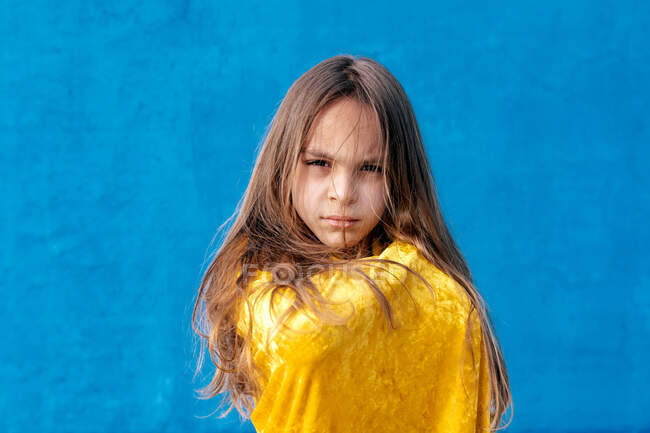 Adolescente serio con el pelo largo envuelto en capa amarilla de pie sobre fondo azul y mirando a la cámara - foto de stock