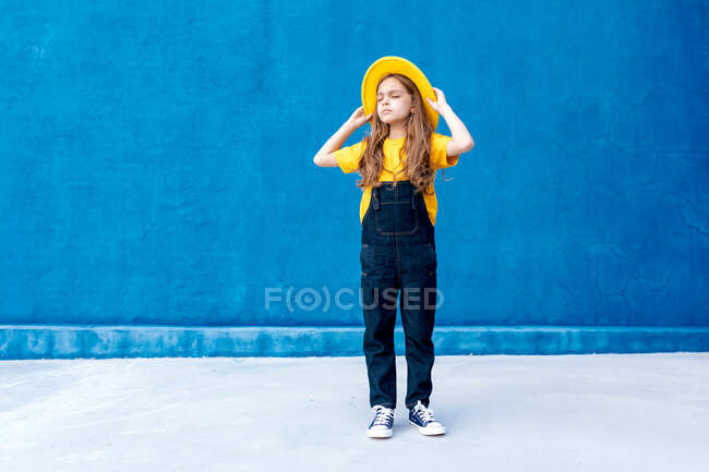Cool pensativo adolescente hipster em macacão e chapéu amarelo de pé com os olhos fechados no fundo da parede azul — Fotografia de Stock