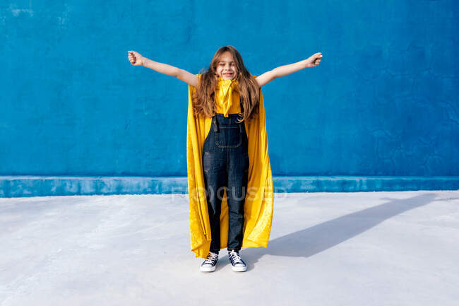 Delighted teenager in superhero yellow cloak standing with outstretched arms with eyes closed on blue background and celebrating victory — Stock Photo