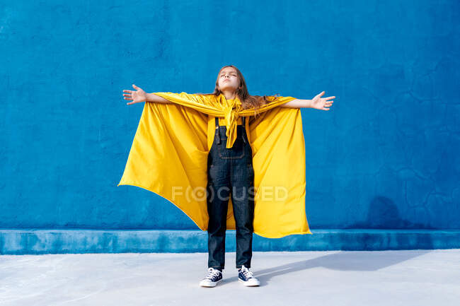Adolescent insensible en manteau jaune superhéros debout avec les bras tendus avec les yeux fermés sur fond bleu et célébrant la victoire — Photo de stock