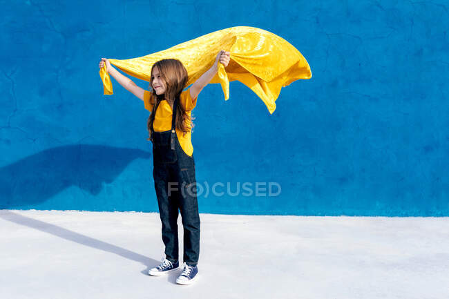 Adolescent ravi en manteau jaune superhéros debout avec les bras tendus regardant loin sur fond bleu et célébrant la victoire — Photo de stock