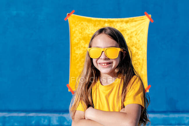 Content teen hipster in sunglasses and with long hair standing with arms crossed on yellow and blue background — Stock Photo