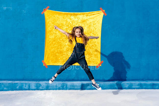 Glücklicher Teenie-Hipster mit langen Haaren springt über den Boden gegen die gelb-blaue Wand und blickt in die Kamera — Stockfoto