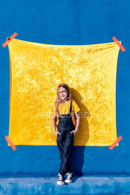 Feliz adolescente hipster com cabelos longos em pé com as mãos no bolso contra a parede amarela e azul e olhando para a câmera — Fotografia de Stock
