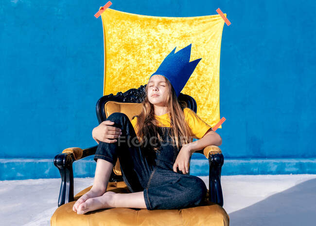 Adolescente pensativo em coroa de papel dourado sentado no trono como rei em fundo azul com os olhos fechados — Fotografia de Stock