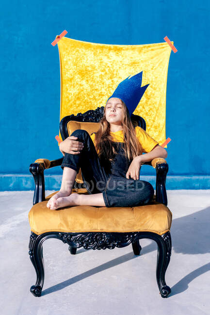 Продуманий підліток у золотій паперовій короні сидить на троні, як король на синьому фоні з закритими очима — стокове фото