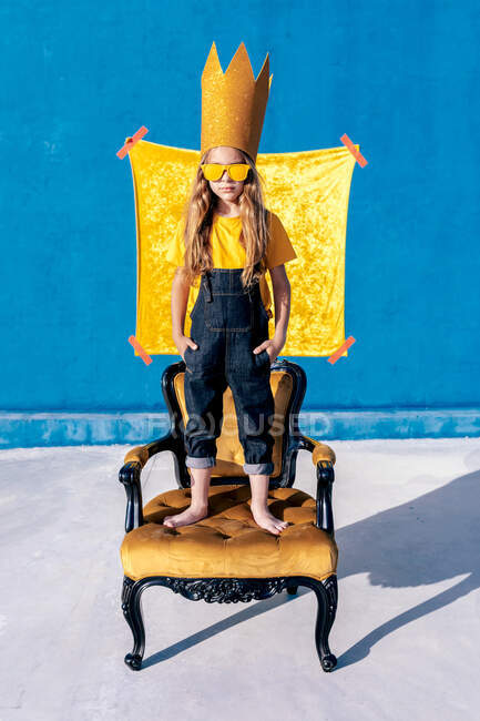 Крутой хипстер-подросток в бумажной короне и солнцезащитных очках стоит на королевском троне и смотрит в камеру, притворяясь королем — стоковое фото