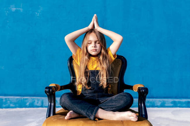 Heiterer Teenie-Hipster sitzt in Lotus-Pose mit geschlossenen Augen und Namaste-Händen und meditiert, während er Yoga auf einem Stuhl auf blauem Hintergrund praktiziert — Stockfoto