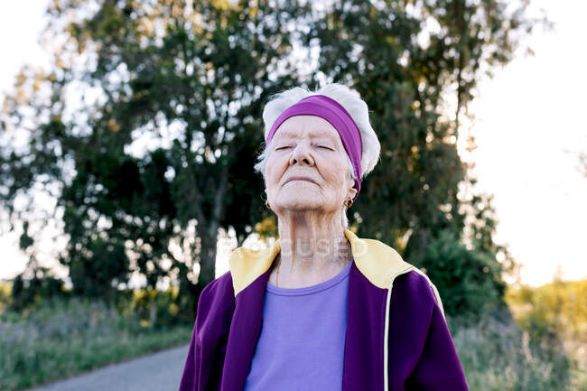 Atleta anciana con los ojos cerrados respirando aire fresco durante el entrenamiento de fitness en el campo - foto de stock