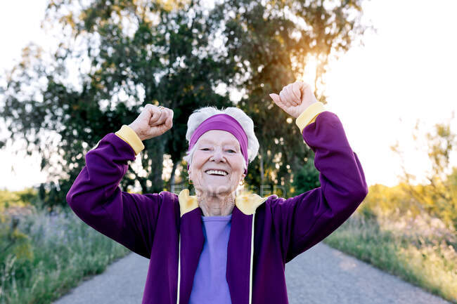 Entzückte Seniorin blickt lächelnd in die Kamera und reckt geballte Fäuste, während sie beim morgendlichen Training in der Natur Erfolge feiert — Stockfoto