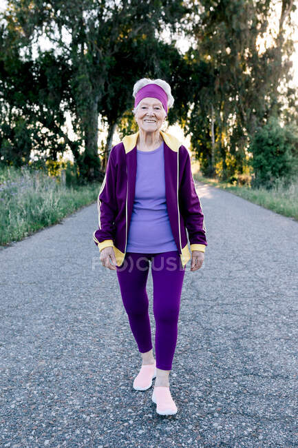 Glad senior female jogger smiling and walking on asphalt road during fitness workout in summer morning in countryside — Stock Photo
