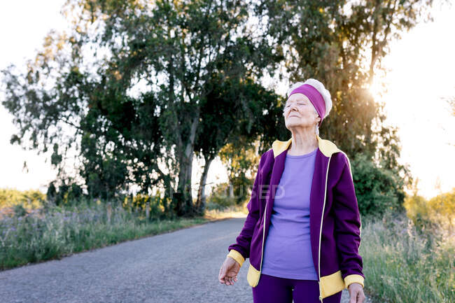 Elderly female athlete with closed eyes breathing fresh air during fitness training in countryside — Stock Photo