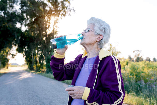 Aged sportswoman sipping fresh water from bottle during break in fitness workout in nature — Stock Photo