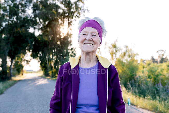 Пенсионерка-бегунья улыбается и ходит по асфальтовой дороге во время фитнес-тренировок летним утром в сельской местности — стоковое фото