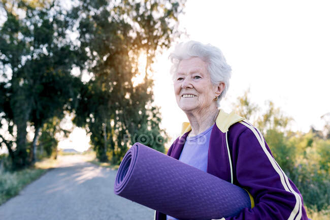 Optimista hembra senior llevando estera enrollada y sonriendo antes de entrenar fitness en la mañana en el campo - foto de stock