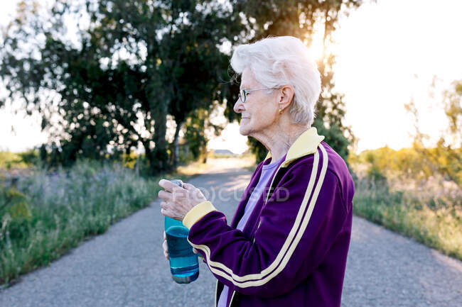 Side view of aged sportswoman drinking fresh water from bottle during break in fitness workout in nature — Stock Photo