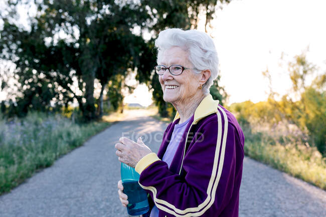 Side view of aged sportswoman drinking fresh water from bottle during break in fitness workout in nature — Stock Photo