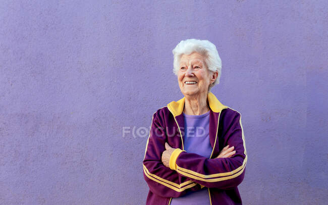 Ehrliche Seniorin in Sportkleidung, die mit verschränkten Armen auf violettem Hintergrund wegschaut — Stockfoto