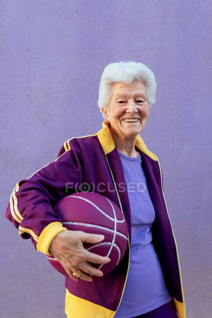 Sonriente jugadora de baloncesto anciana con pelo gris en ropa deportiva mirando a la cámara sobre fondo violeta - foto de stock