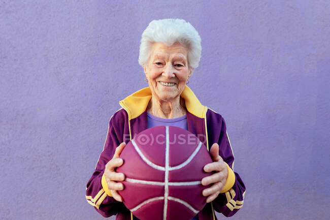 Усміхнена літня баскетболістка з сірим волоссям у спортивному одязі дивиться на камеру на фіолетовому фоні — стокове фото
