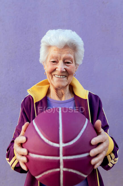 Sorridente anziano giocatore di basket femminile con i capelli grigi in abiti sportivi guardando la fotocamera su sfondo viola — Foto stock