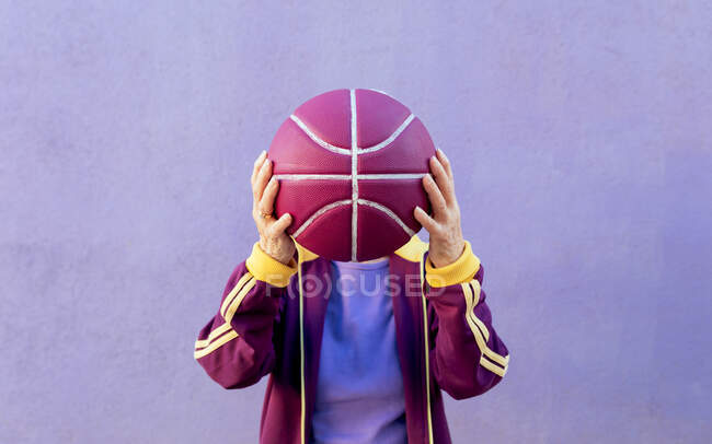 Anonimo senior giocatore di basket femminile in abbigliamento sportivo che copre il viso con la palla su sfondo viola — Foto stock