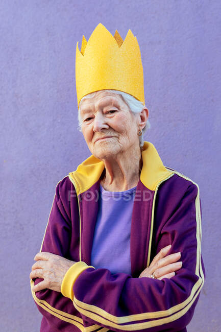 Selbstbewusste ältere Athletin in Sportbekleidung und dekorativer Krone blickt mit verschränkten Armen auf lila Hintergrund in die Kamera — Stockfoto