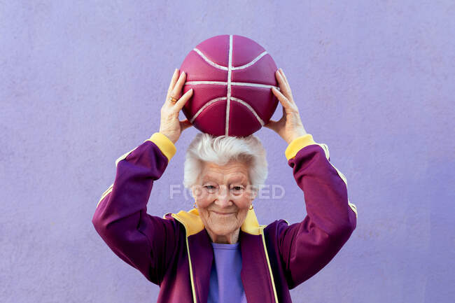Усміхнена літня баскетболістка з сірим волоссям у спортивному одязі дивиться на камеру на фіолетовому фоні — стокове фото