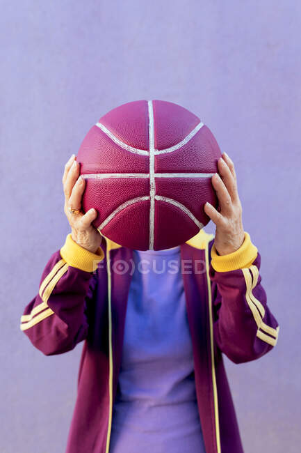 Jugadora senior de baloncesto anónima en ropa deportiva cubriendo la cara con pelota sobre fondo púrpura - foto de stock