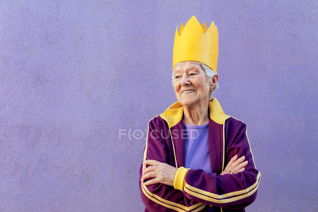 Fiduciosa atleta donna anziana in abbigliamento sportivo e corona decorativa guardando lontano con le braccia incrociate su sfondo viola — Foto stock