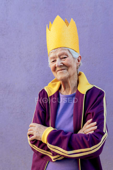 Atleta femenina segura de edad avanzada en ropa deportiva y corona decorativa mirando a la cámara con los brazos cruzados sobre fondo púrpura - foto de stock