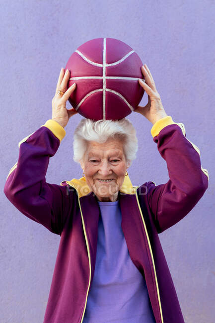 Lächelnde ältere Basketballspielerin mit grauen Haaren in Sportkleidung blickt in die Kamera auf violettem Hintergrund — Stockfoto
