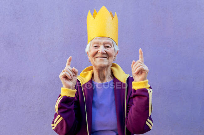 Content senior female athlete in sportswear and paper crown pointing up with fingers while looking at camera on violet background — Stock Photo